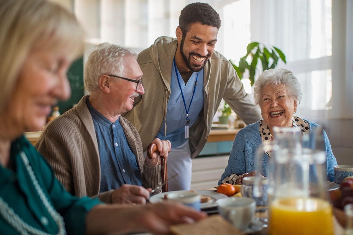 Residents at The Commons enjoy a lively social gathering, sharing a meal and laughter with a caregiver in a warm, welcoming environment.